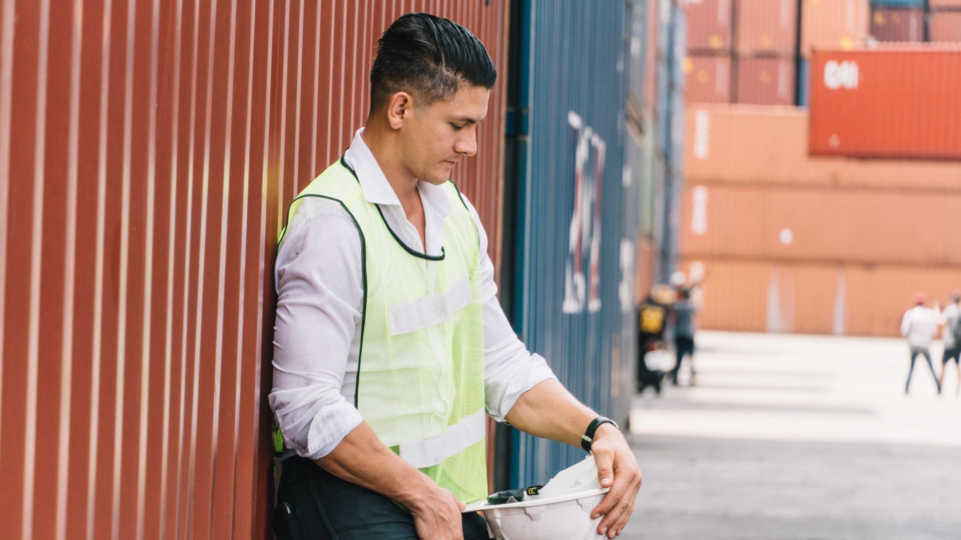 Worker with hardhat leaning despondently against a fence outside a factory